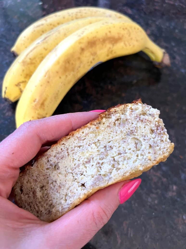 Melanie Mendelson from melaniecooks.com, holding a slice of almond flour banana bread with ripe bananas in the background, showing soft and moist texture.