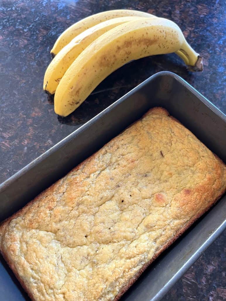 Gluten-free almond flour banana bread cooling in loaf pan next to fresh bananas on kitchen counter.