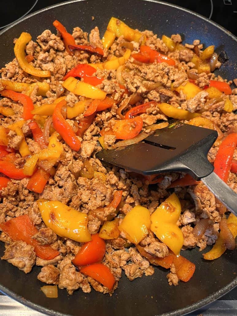 Ground turkey fajitas being stirred in a skillet with a spatula, showing the step of mixing in the peppers and onions.