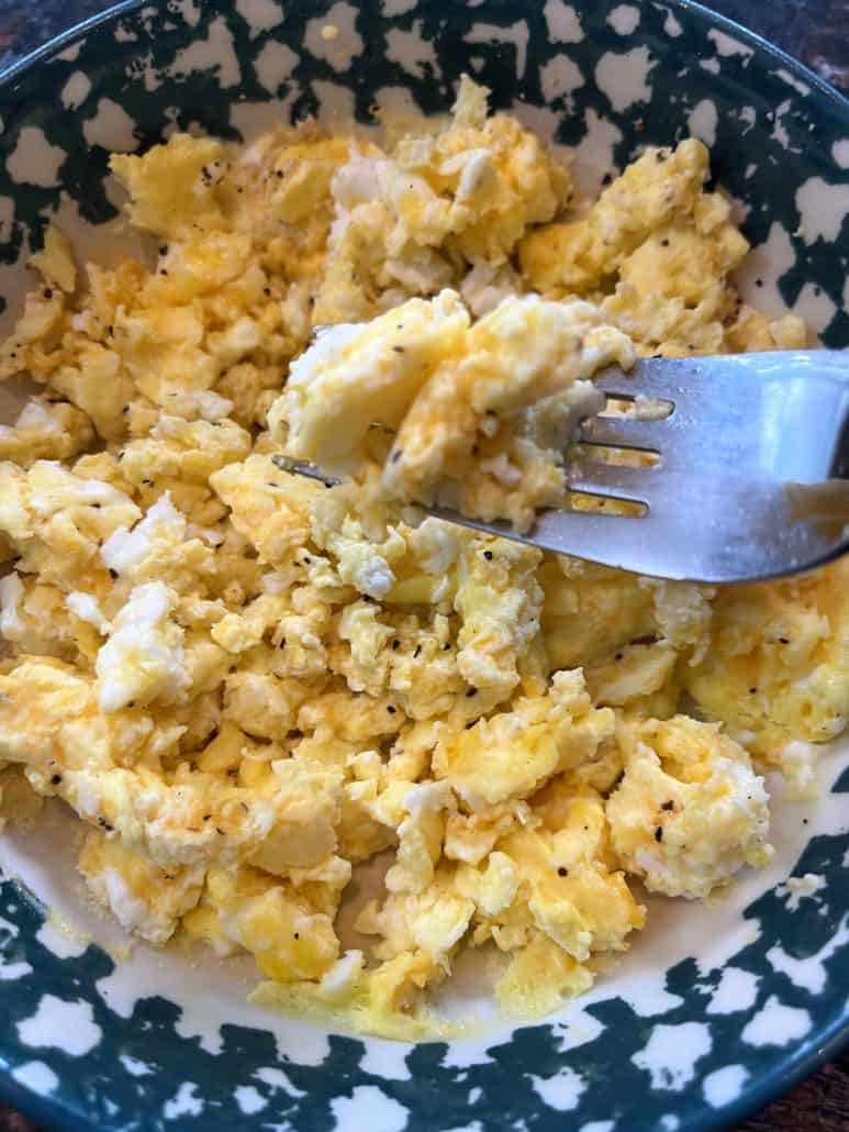 Close-up of a fork lifting fluffy microwave scrambled eggs from a patterned bowl, showing the soft texture.