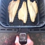 Three frozen branzino fillets cooking in an air fryer basket as shown in the Air Fryer Branzino Fillets recipe.