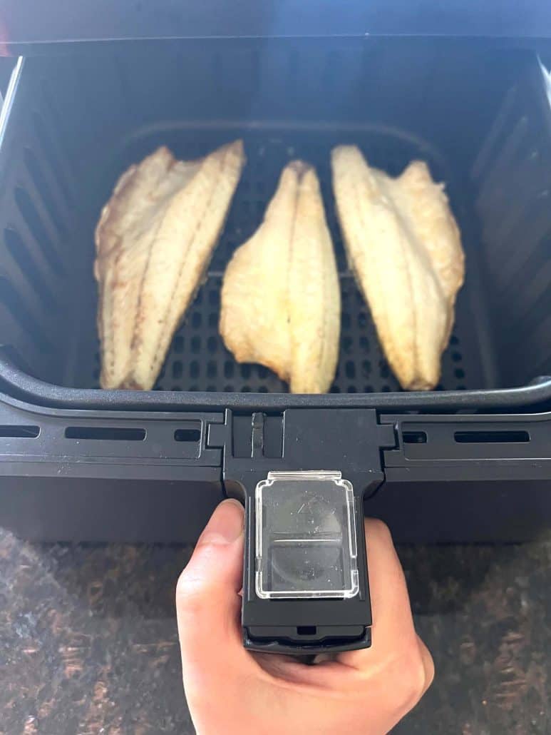Hand pulling out air fryer basket with three cooked branzino fillets, as shown in the Air Fryer Branzino Fillets recipe.