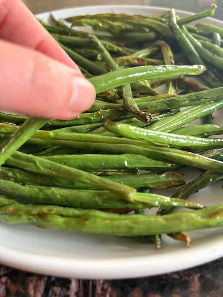 Hand picking up a crispy roasted French green bean (Haricot Vert) from a plate, showing the final result.
