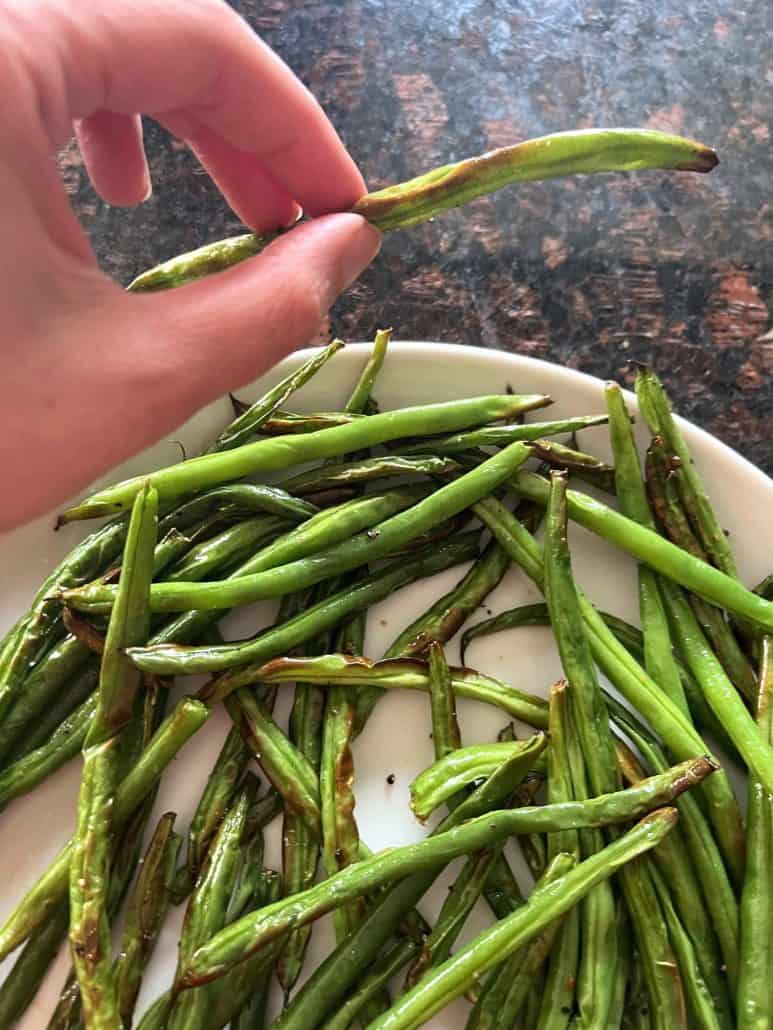 Hand holding a single roasted French green bean (Haricot Vert) above a plate of air-fried green beans, ready to enjoy.