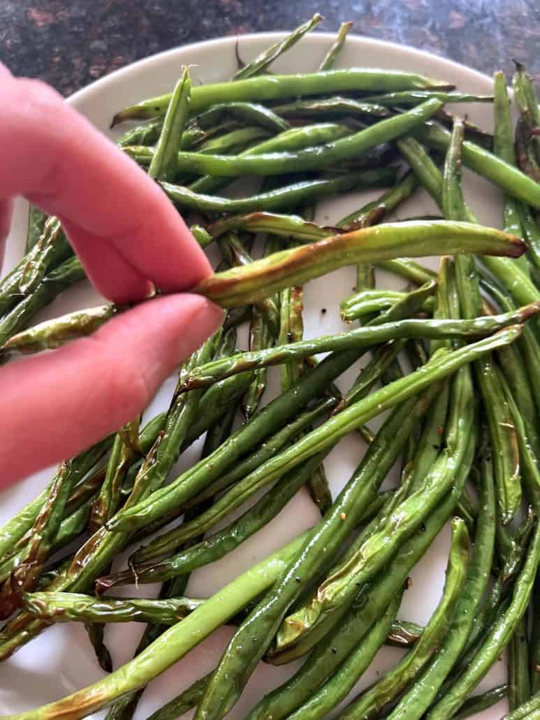 Hand holding a crispy air-fried French green bean (Haricot Vert) above a plate of freshly cooked green beans from the air fryer.