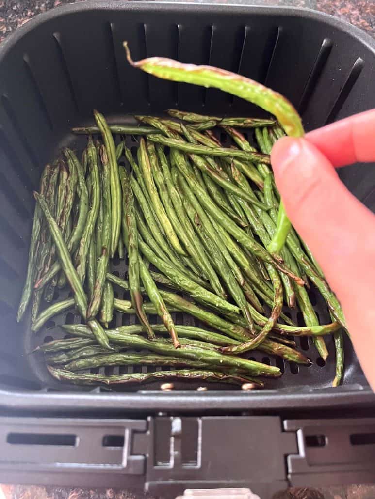 Hand holding a perfectly roasted French green bean (Haricot Vert) above the air fryer basket, showing the crispy texture.
