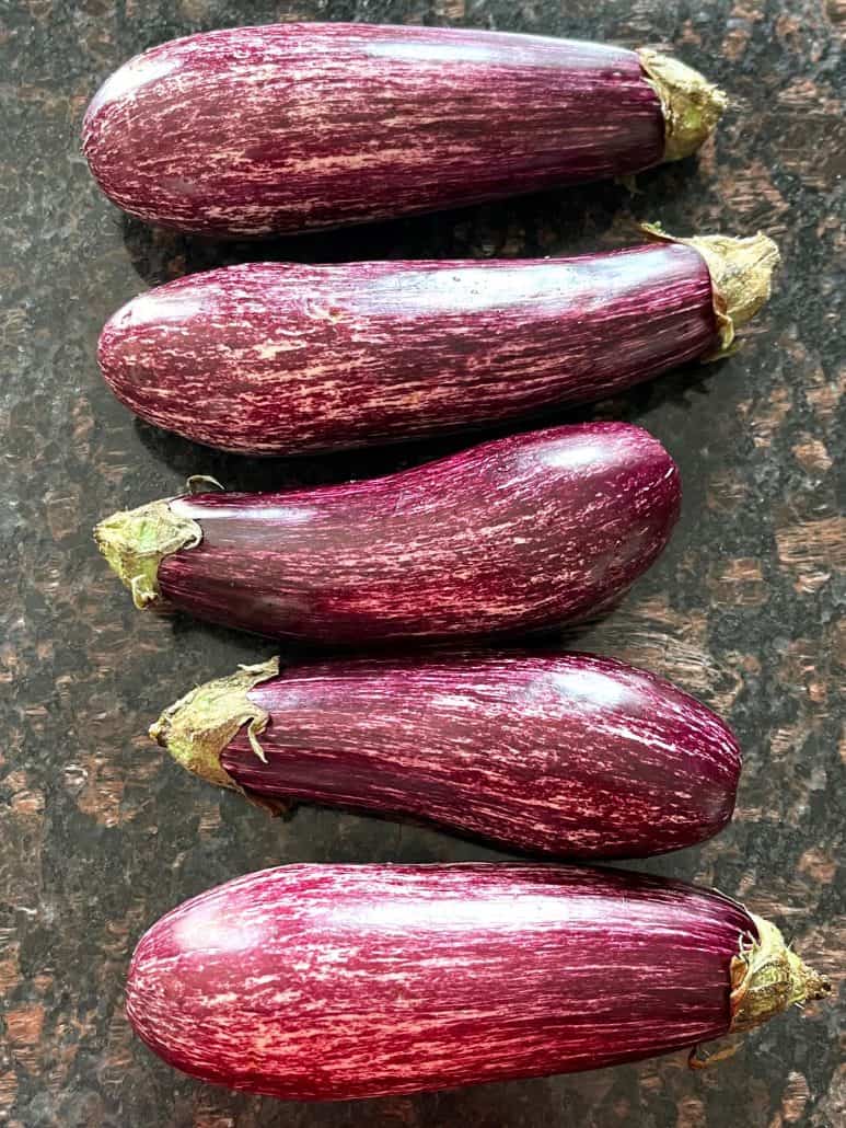 Five raw graffiti eggplants are lined up on the countertop, prepped and ready to be chopped.