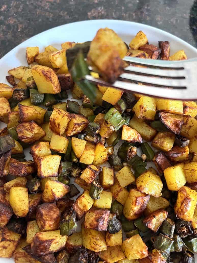 Okra and potatoes lifted on a fork above a plate, showcasing the crispy, golden texture of this easy veggie dish.