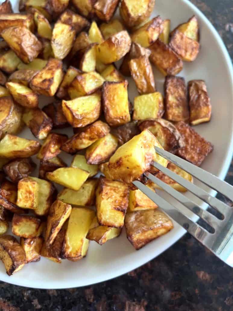 Close-up of a fork lifting a crispy air fryer potato cube from a plate full of golden skin-on potatoes.