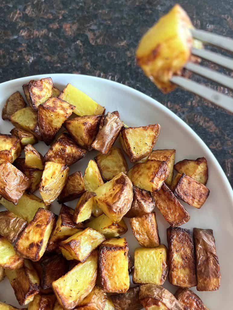 A fork holds up a single crispy, golden air-fried potato cube above a plate full of roasted skin-on potatoes.