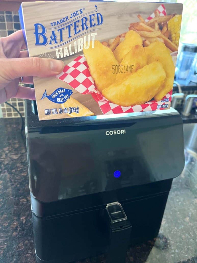 Holding a box of Trader Joe’s Battered Halibut in front of a black air fryer, ready to cook the fillets.