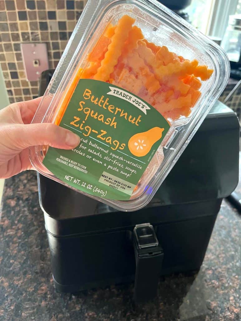 Holding a package of Trader Joe’s Butternut Squash Zig-Zags in front of an air fryer.
