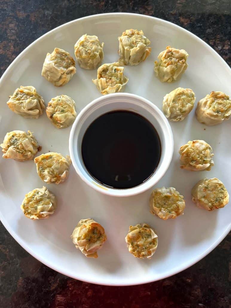 Air-fried Trader Joe’s Chicken Shu Mai dumplings arranged in a circle on a white plate with a cup of dipping sauce in the center, ready to serve.