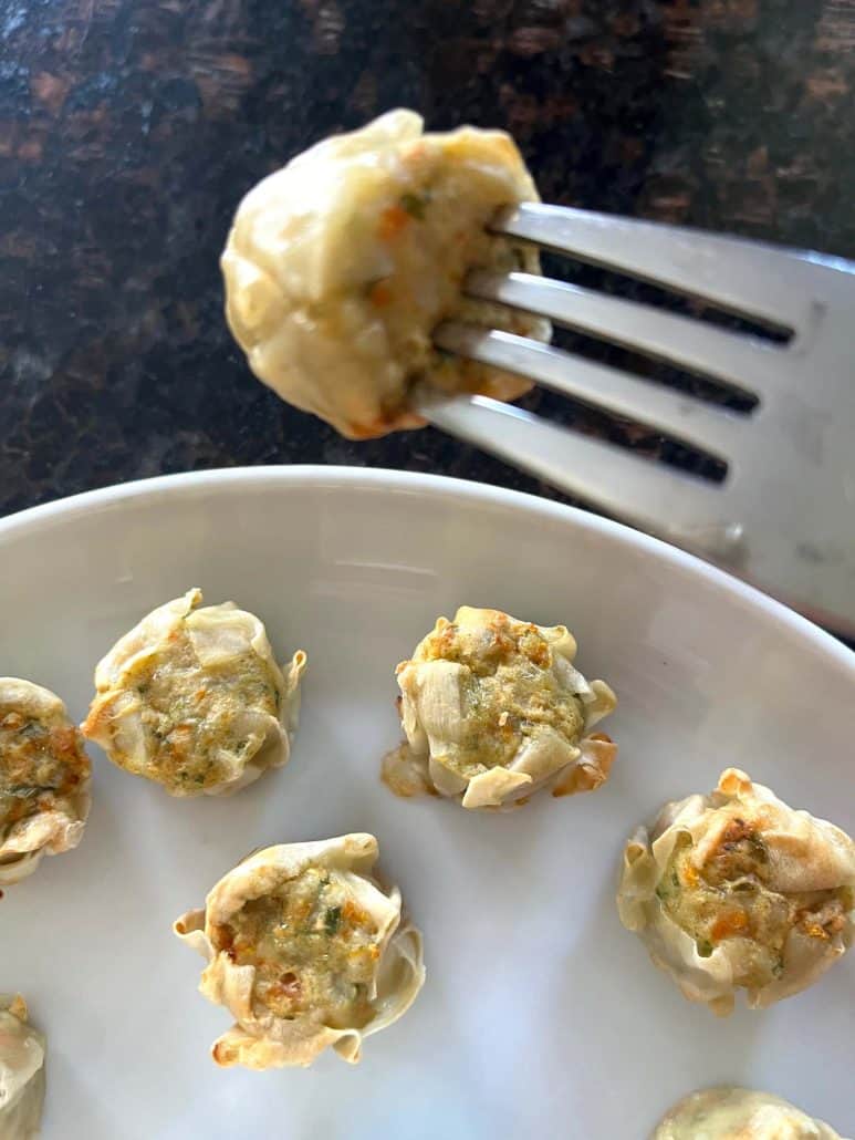 Close-up of a Trader Joe’s Chicken Shu Mai dumpling held on a fork above a plate with more cooked dumplings.
