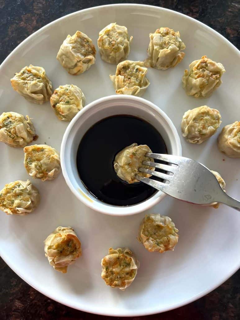 Fork dipping a Trader Joe’s Chicken Shu Mai dumpling into soy-based sauce, with the rest of the dumplings arranged around the dipping cup on a white plate.