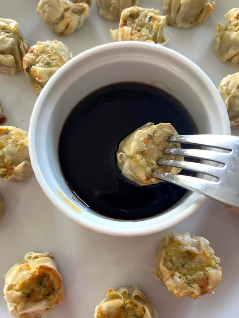 Fork holding a Trader Joe’s Chicken Shu Mai dumpling over a small bowl of dipping sauce, surrounded by more dumplings on a white plate.