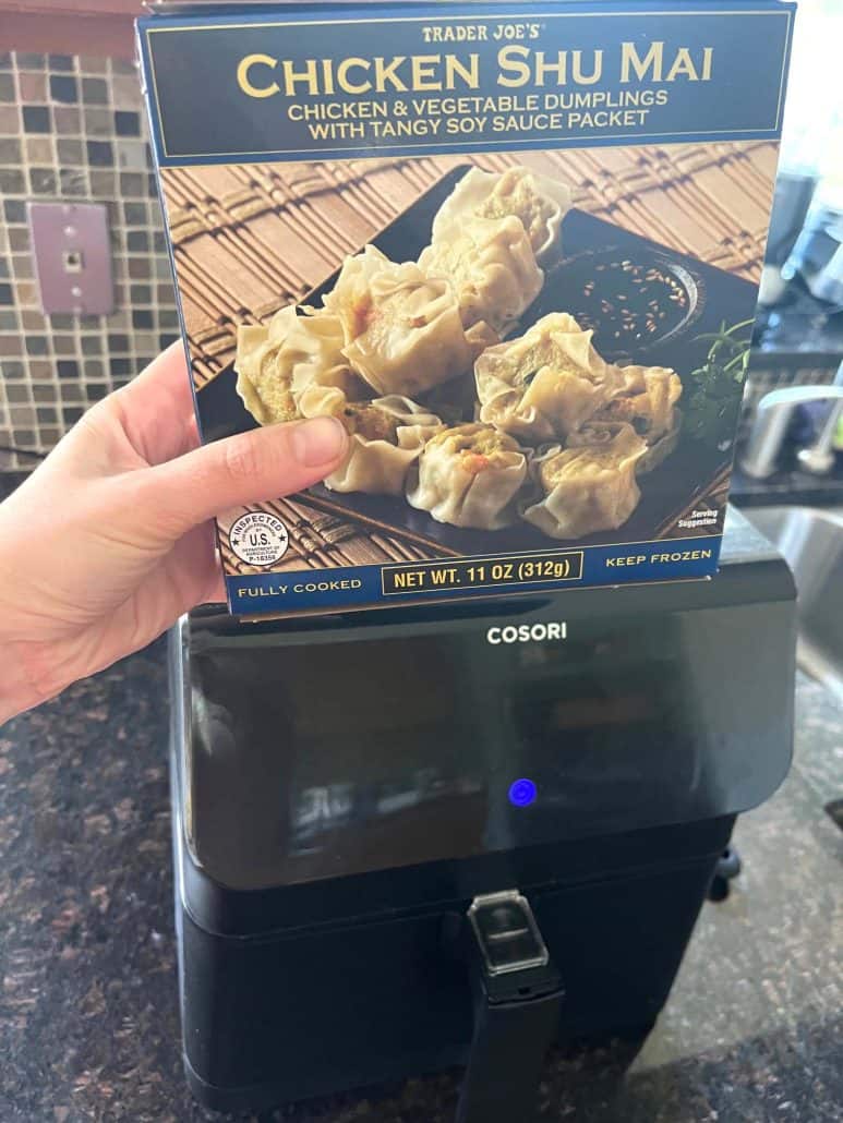 Holding a box of Trader Joe’s Chicken Shu Mai dumplings in front of an air fryer, showing the product.