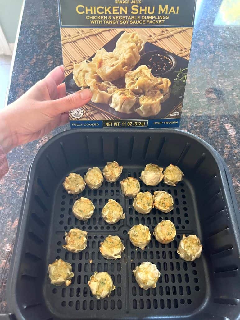 Holding a box of Trader Joe’s Chicken Shu Mai over an air fryer basket filled with the frozen dumplings arranged in a single layer, ready to cook using the steam method.