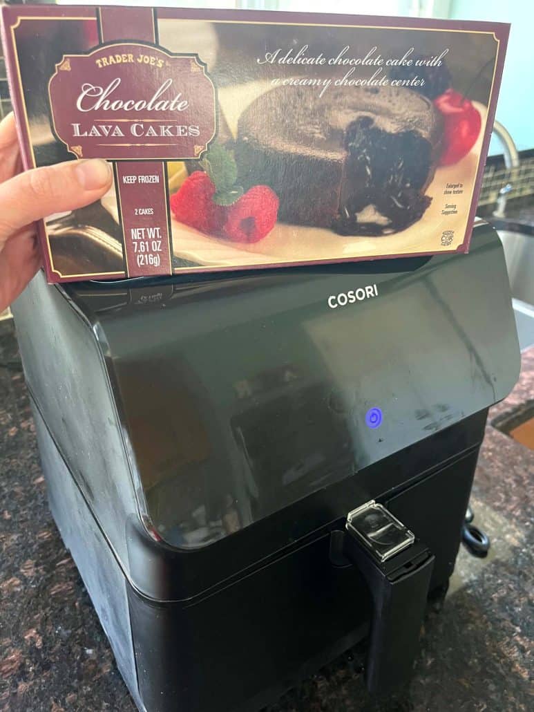 Holding a box of Trader Joe’s Chocolate Lava Cakes in front of an air fryer, ready to cook the frozen dessert using the air fryer method.