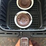 Two Trader Joe’s chocolate lava cakes cooking in an air fryer basket, showing how they are baked directly in their oven-safe cups from frozen.