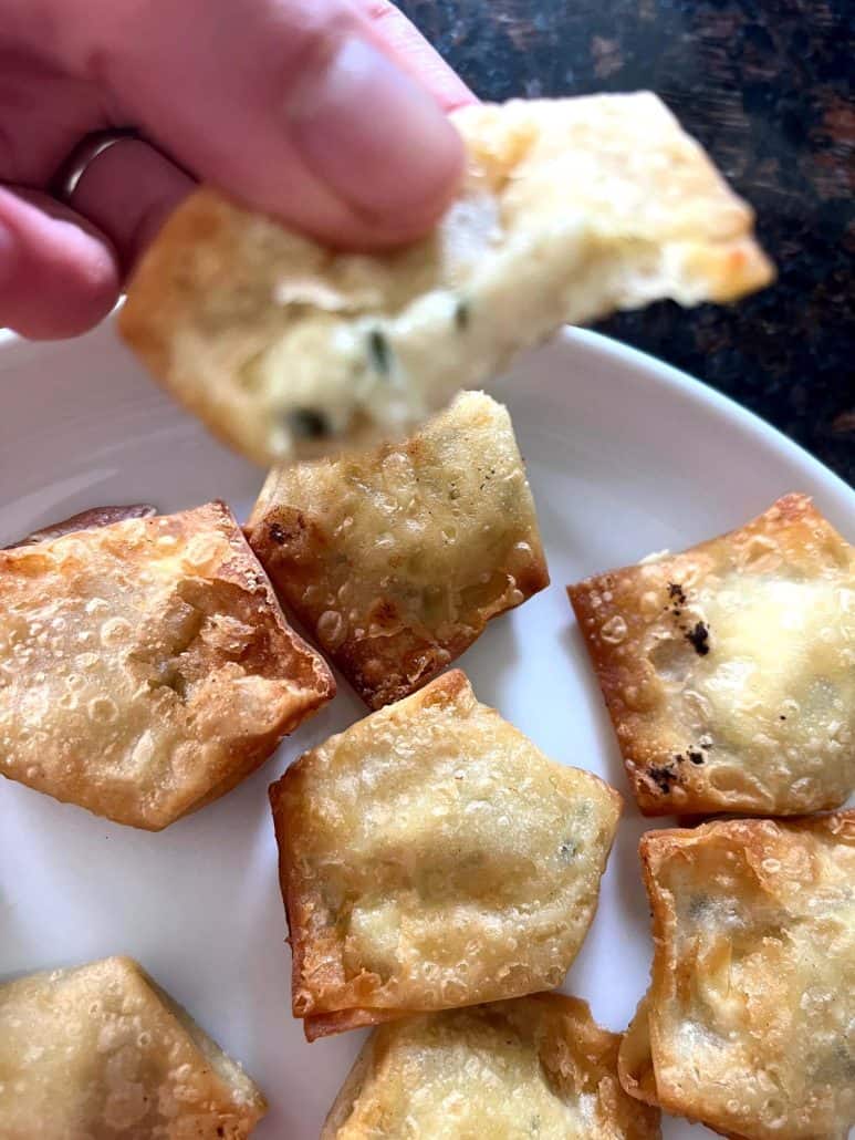 Hand holding a bitten air-fried Trader Joe’s wonton, revealing the melted jalapeño cream cheese filling, with more crispy wontons on a plate below.