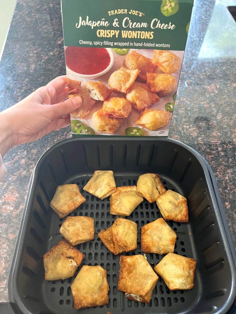 Hand holding the box of Trader Joe’s Jalapeño & Cream Cheese Crispy Wontons above freshly air-fried wontons in the basket, showcasing the before-and-after from the easy air fryer recipe.