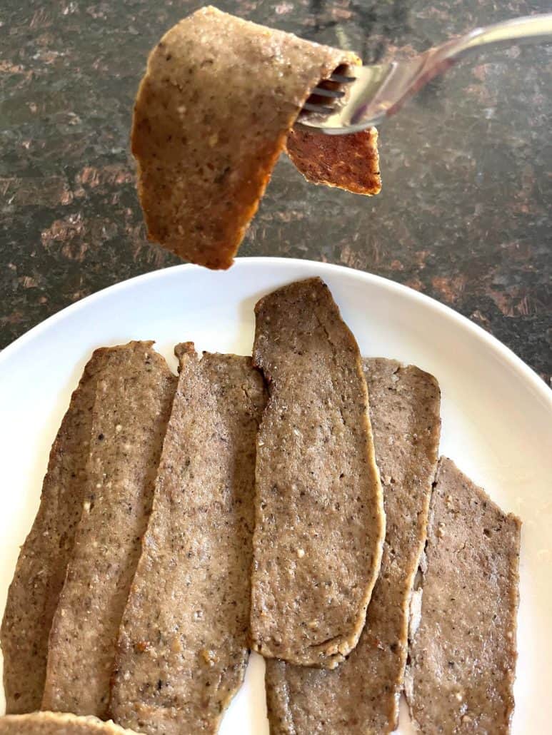Fork holding up a cooked slice of Trader Joe’s gyro meat showing crispy browned edge, with more slices on a plate below.