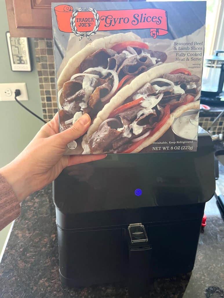 Hand holding a box of Trader Joe’s Gyro Slices in front of an air fryer, ready to cook for a quick meal