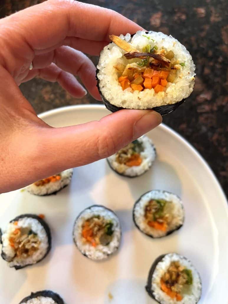 Melanie Mendelson from melaniecooks.com holding a piece of cooked Trader Joe's kimbap, showing the filling, with the rest of the kimbap on a plate.