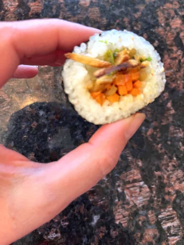 Melanie Mendelson from melaniecooks.com holding a piece of Trader Joe's kimbap, showing the filling inside with a close-up view of the rice and vegetables.