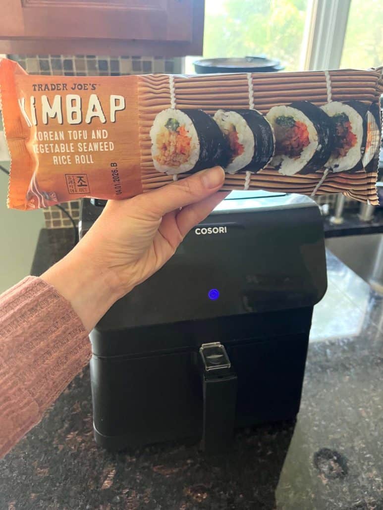 Melanie Mendelson from melaniecooks.com holding a package of Trader Joe's frozen kimbap in front of an air fryer, ready for cooking.
