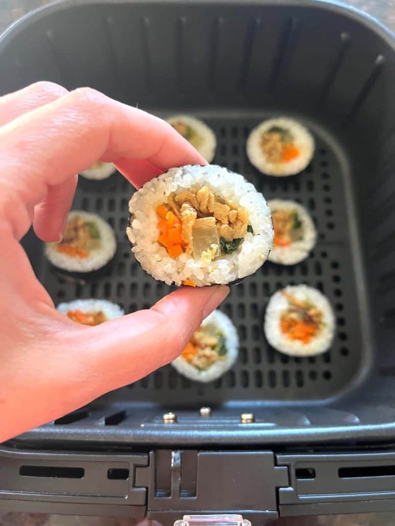 Melanie Mendelson from melaniecooks.com holding a piece of Trader Joe's kimbap, showing the filling, with the rest of the kimbap arranged in the air fryer basket.