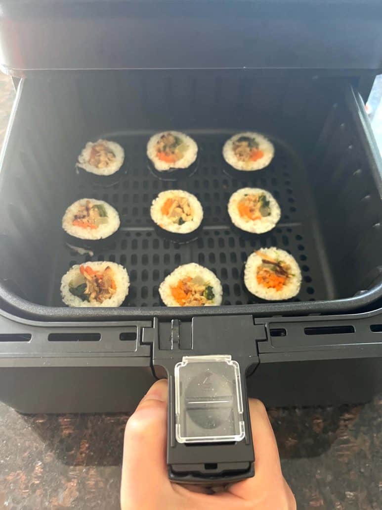 Melanie Mendelson from melaniecooks.com holding an air fryer basket with Trader Joe's frozen kimbap arranged inside, ready to cook.