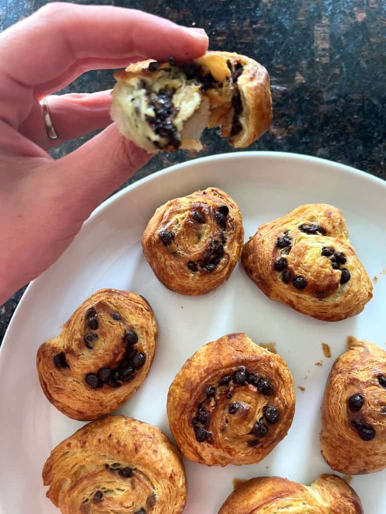 Hand holding a Trader Joe’s Mini Croissant, broken in half to reveal the melted chocolate filling, with the rest of the croissants on the plate.