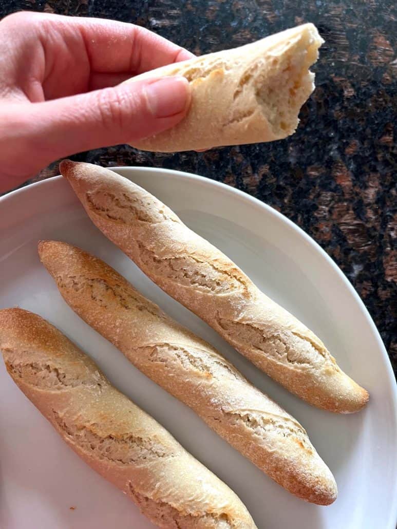 Hand holding a piece of warm, air-fried Trader Joe’s mini French baguette with more cooked baguettes on a plate below, showing the soft inside and crispy crust.