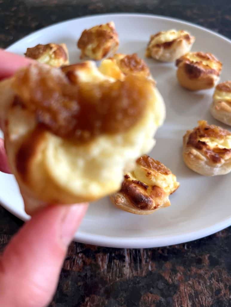 Close-up of a cheesy Trader Joe’s Pastry Bite held in hand, showing caramelized onion filling with more bites on a white plate in the background.