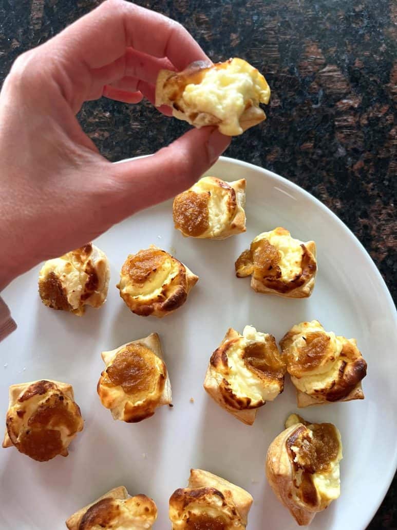 Hand holding a Trader Joe’s Pastry Bite showing the melted cheese filling, with more golden brown bites arranged on a white plate below.