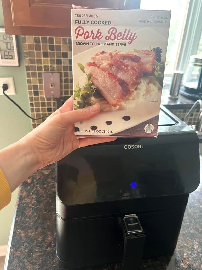 Holding a box of Trader Joe’s Fully Cooked Pork Belly in front of an air fryer—ready to make the quick and crispy air fryer pork belly recipe.