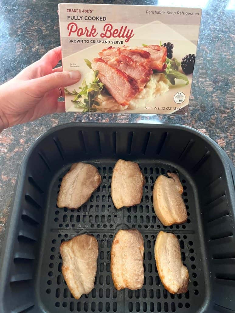 Holding a box of Trader Joe’s Fully Cooked Pork Belly above an air fryer basket filled with sliced pork belly.