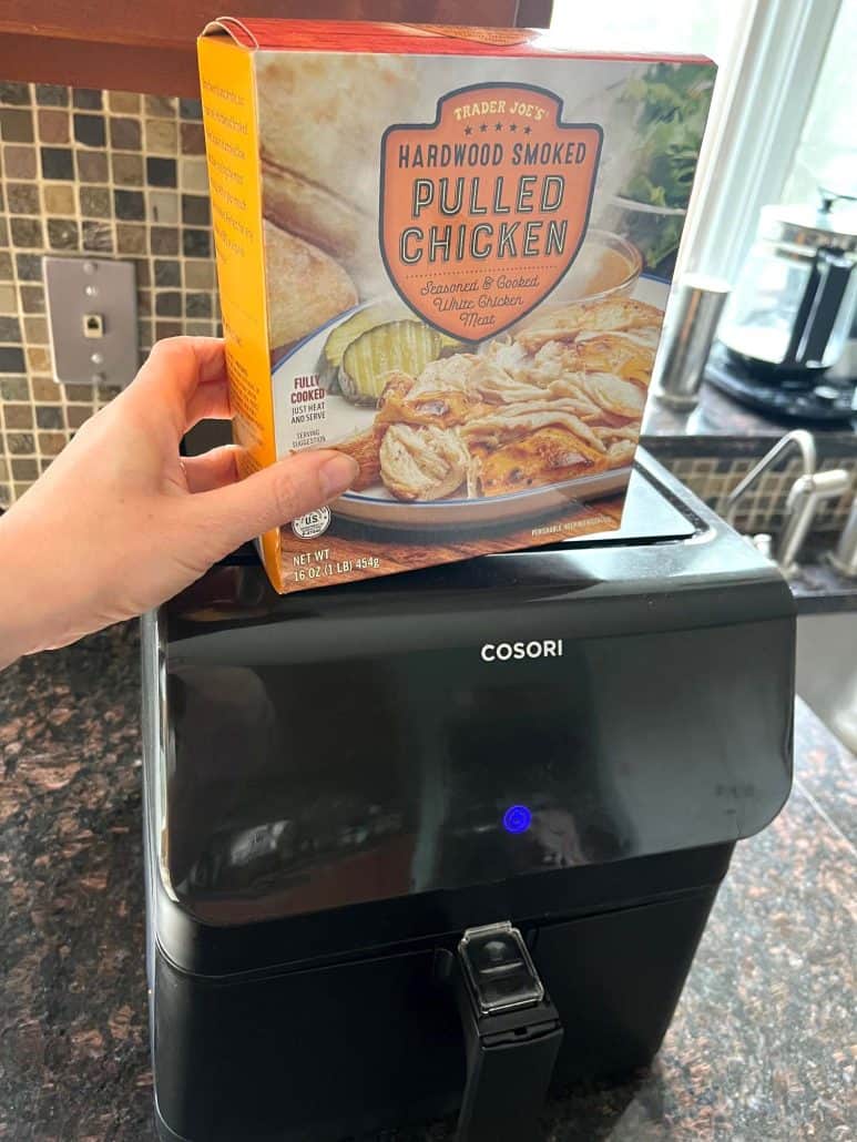 Hand holding a package of Trader Joe’s Pulled Chicken in front of an air fryer.