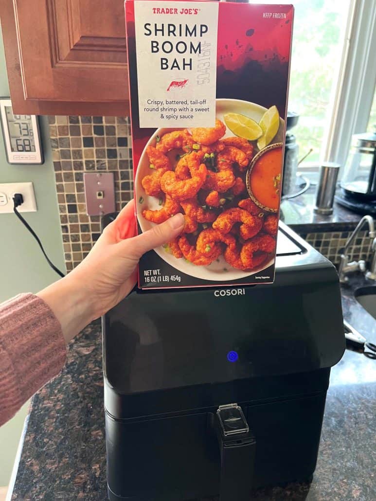 Holding a box of Trader Joe’s Shrimp Boom Bah in front of an air fryer, ready to make the crispy shrimp.