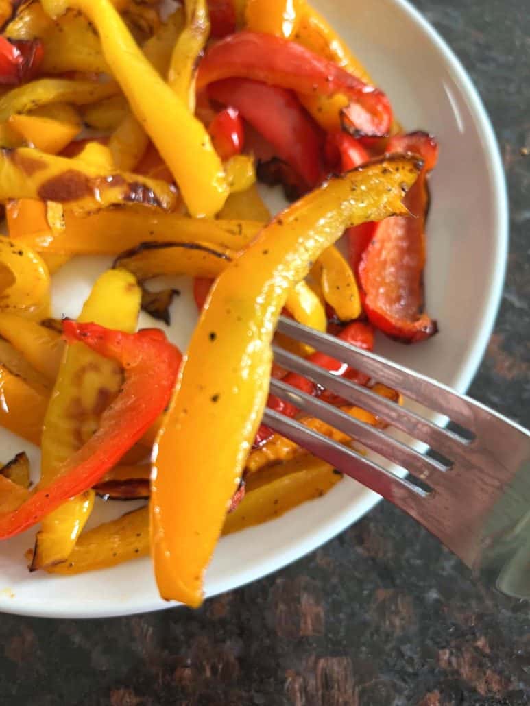 Close-up of a fork holding a roasted orange bell pepper slice over a plate of tri-color peppers—showcasing the tender texture.