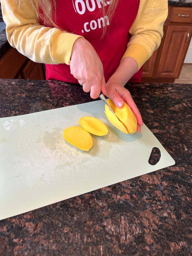 Cutting the second cheek from a peeled Philippine Carabao (Manila) mango on a cutting board, continuing the process of slicing around the pit.