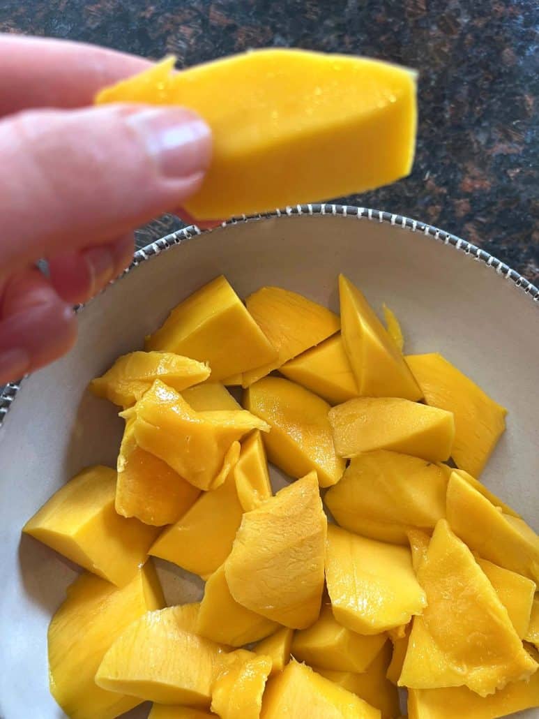 Hand holding a slice of Philippine Carabao (Manila) mango above a bowl filled with freshly cut mango pieces, showcasing the final result of the cutting process.