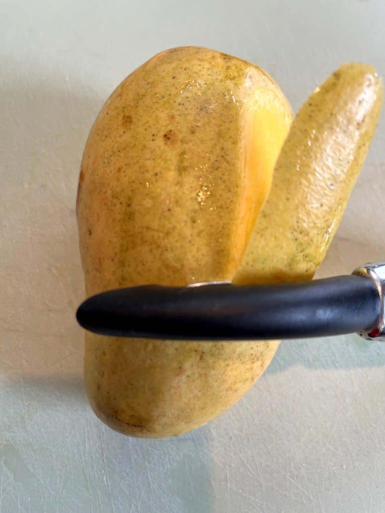 Peeling a Philippine Carabao (Manila) mango with a vegetable peeler, demonstrating the first step in the mango cutting process.