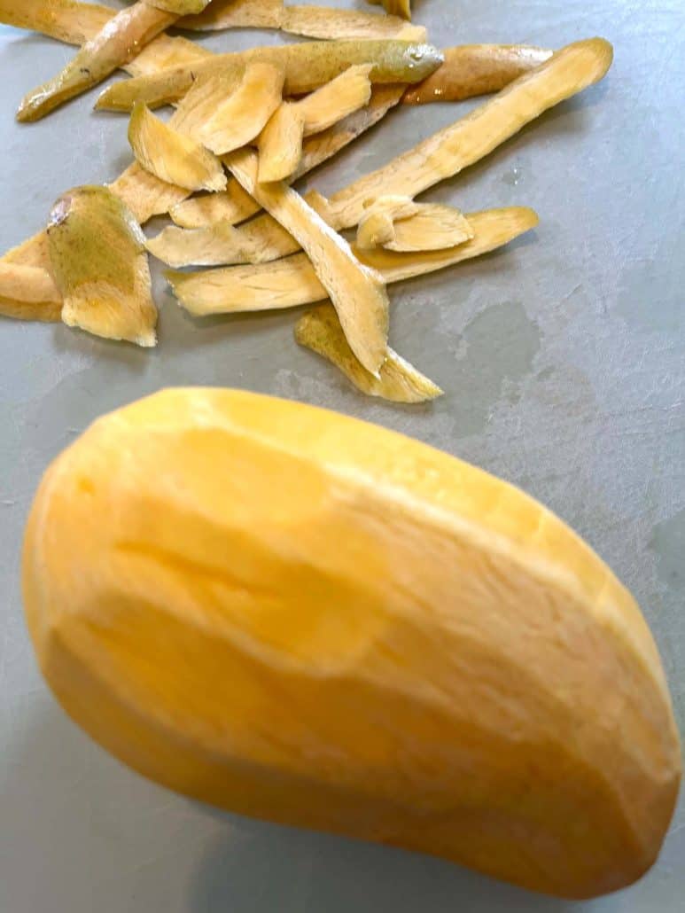 Fully peeled Philippine Carabao (Manila) mango with skin scraps in the background, ready for slicing in the next step of the tutorial.