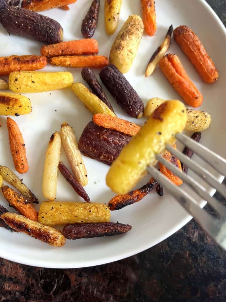 Fork holding a roasted yellow baby carrot above a plate of colorful tri-color carrots, showing tenderness.