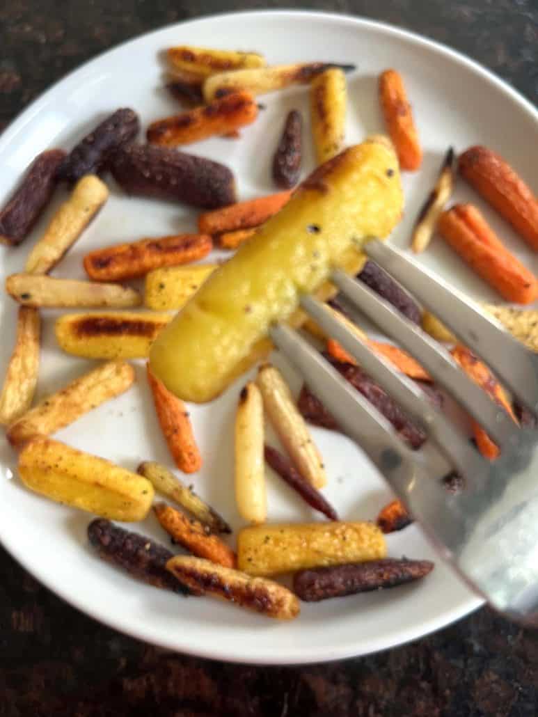 Fork holding a tender roasted yellow carrot above a plate of tri-color baby carrots, showcasing doneness from the Oven Roasted Tri-Color Carrots Recipe.