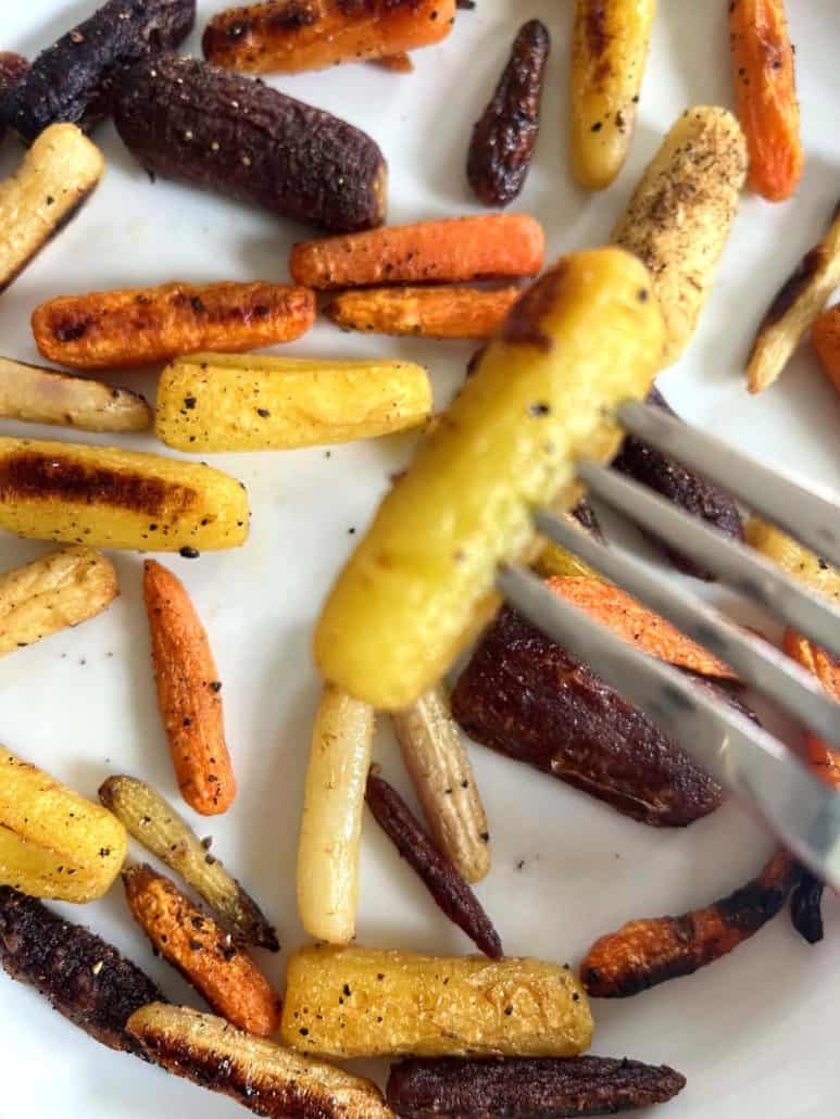 Close-up of a fork piercing a roasted yellow carrot over a plate of tri-color baby carrots, illustrating the fork test step.