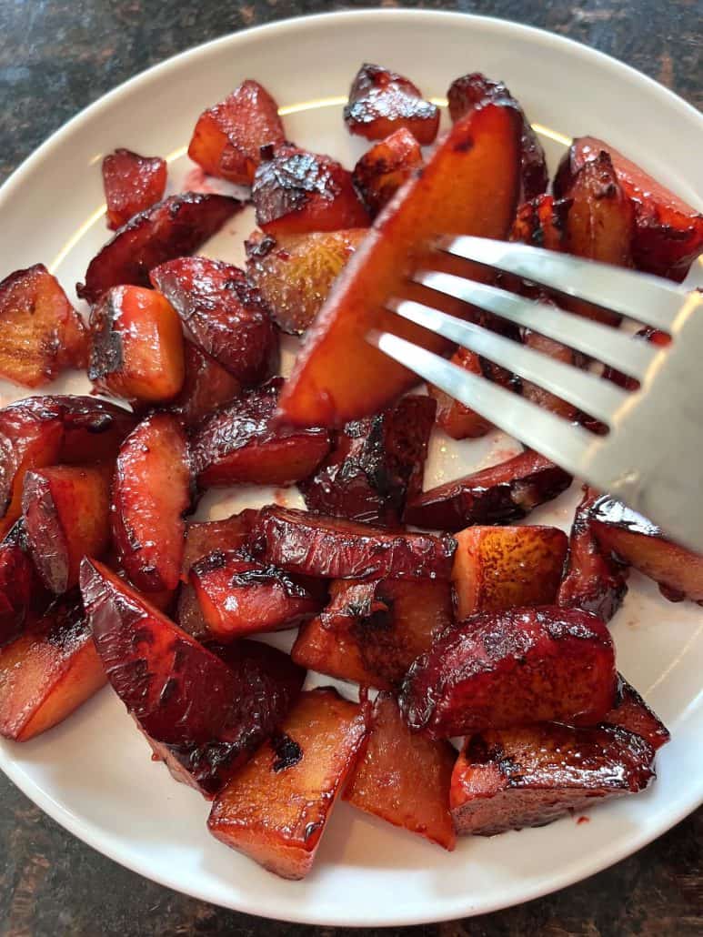 Fork lifting a caramelized plum slice from a plate of sauteed plums, highlighting the tender texture and rich color of the finished dish.